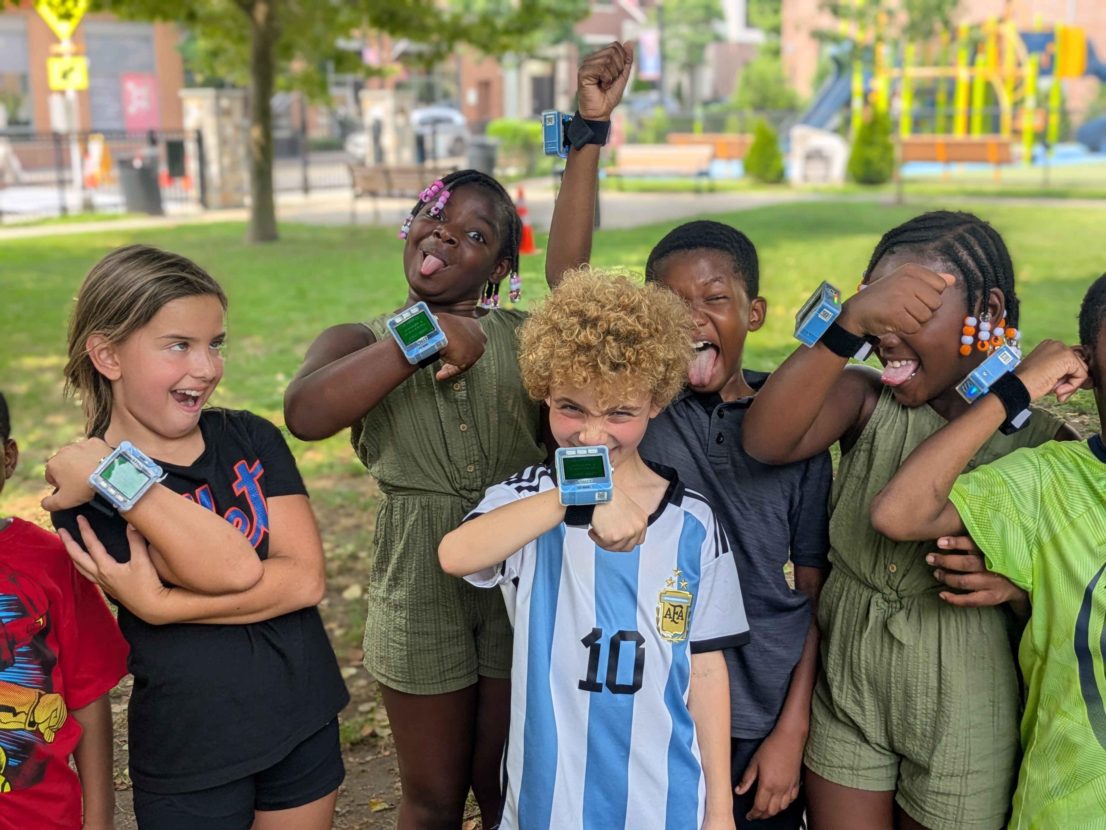 A group of children holding their hands up, highlighting ZTAG watches on their wrists. There are a group of girls in the background and a boy with an argentina soccer jersey in the front.