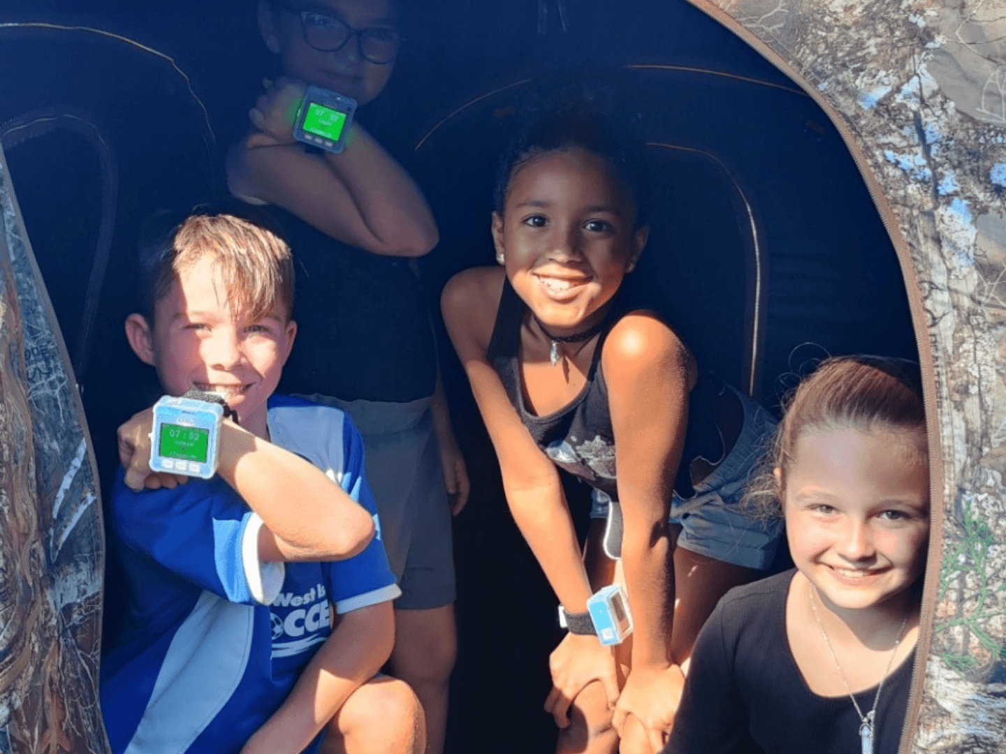 a group of children wielding ztag devices on their wrists. they are smiling at the camera and posing in different positions. the children are inside a hunting popup tent