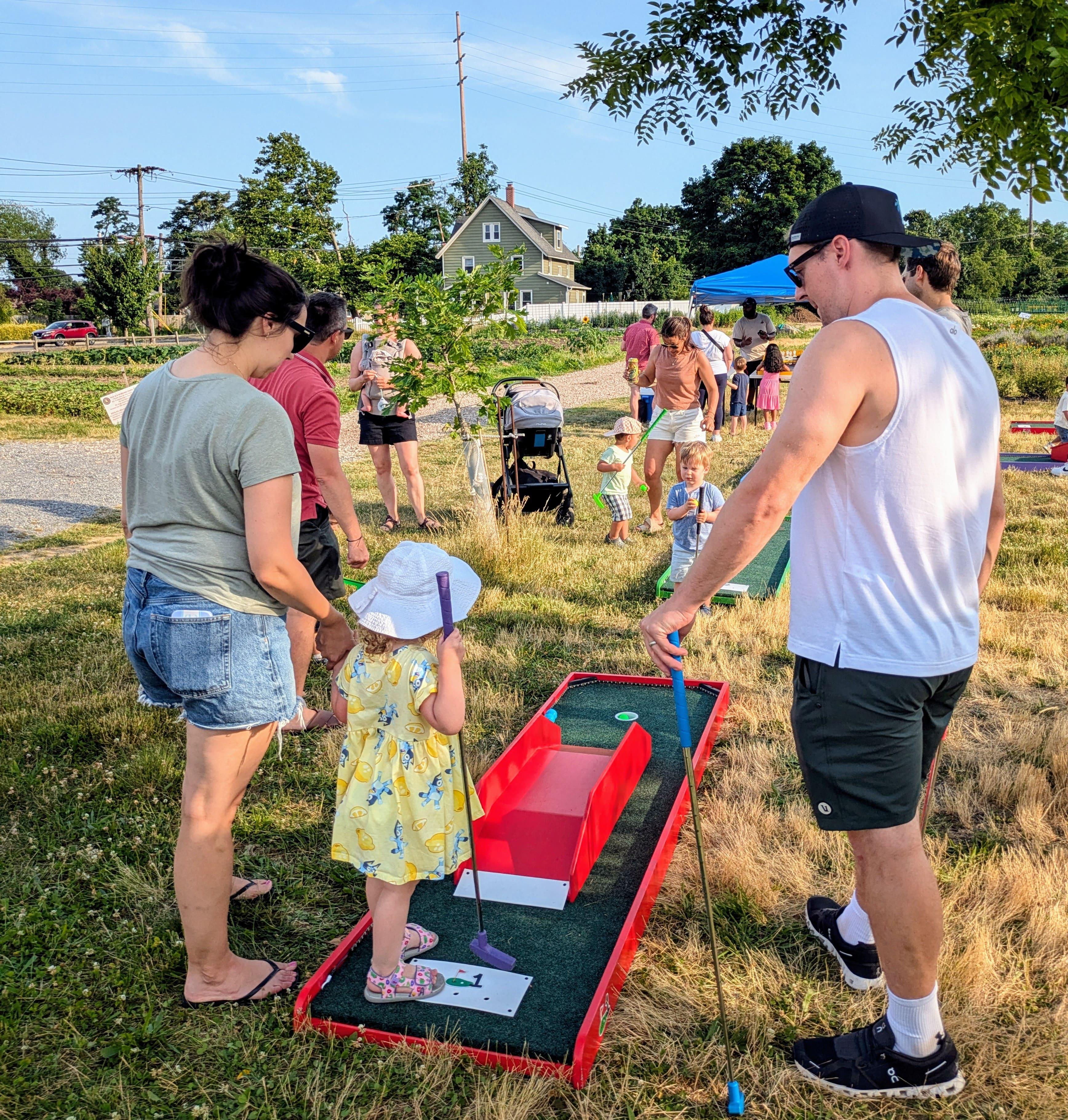 A family standing on a mobile mini golf course. The course had LED lights and there are a lot of people around watching the family putt.